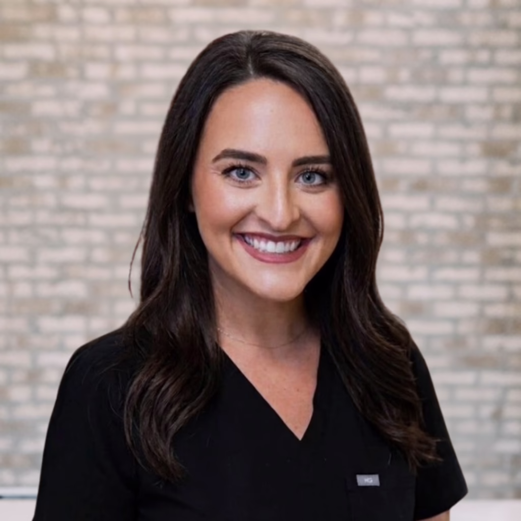 Smiling woman with dark hair in black scrubs, posed against a brick wall backdrop.